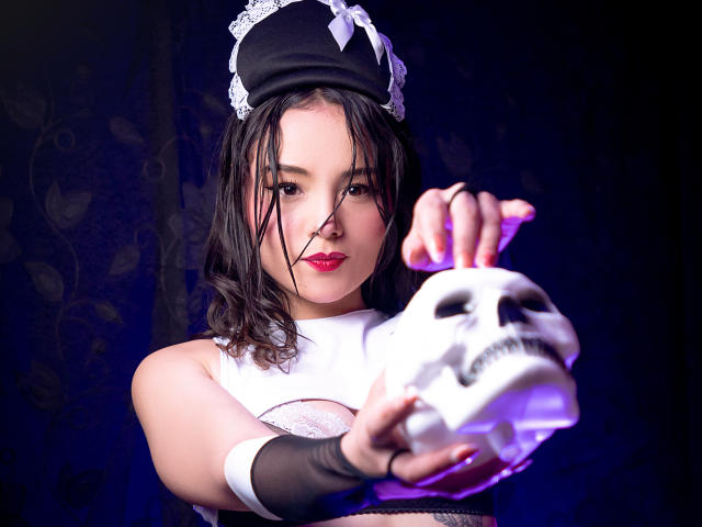 ViolettaMidnight mit nassen, schwarzen Haaren, roten Lippen und einem weißen Spitzenstirnband, die nach vorne reicht und eine leuchtend violette Hand hat. Schwarzer Spitzenhandschuh am rechten Arm, intensiver Blick, dunkler Hintergrund. Extrem intensiv..