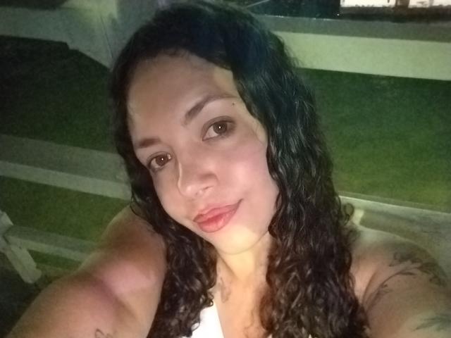 Foto em close-up de RubbyStrongX com cabelos pretos longos e ondulados, pele marrom clara e lábios cheios. RubbyStrongX tem uma expressão relaxada, a cabeça ligeiramente inclinada e o peito superior visível. O fundo é escuro e desfocado..