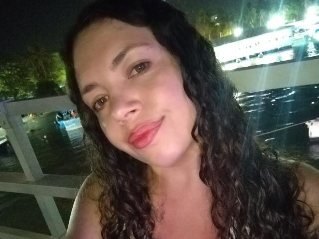 Close-up of RubbyStrongX with curly black hair, light brown skin, and red lipstick. She's smiling slightly, looking at the camera with brown eyes. Bright lights in the background. Fucking sexy.