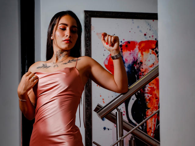 SamantaAbby, gekleed in een strak, glanzend roze jurkje, staat met één hand op haar borst en de andere hand omhoog, waarbij ze tattoos op haar schouders. Kunstwerk met rode en zwarte spatten op de achtergrond.