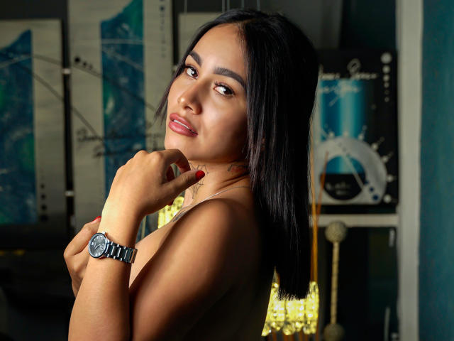 Foto van SamantaAbby met lang zwart haar, die over haar schouder kijkt, met haar hand in de buurt van haar mond, en een zilveren horloge draagt. toploos, achtergrond in een helder geel, donkere, sfeervolle belichting, sexy, verleidelijke pose.