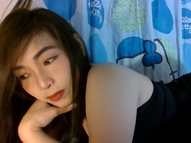 SaigonLady69, mit langem, schwarzem Haar und rotem Lippenstift, lehnt sich auf ihre Hand und blickt verführerisch. Sie trägt ein schwarzes, trägerloses Oberteil vor blauen und gelben Vorhängen mit Muster. Verdammt sexy Atmosphäre..