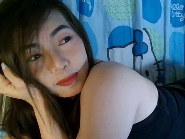 Close-up van SaigonLady69, een Aziatische vrouw met lang zwart haar, rode lippenstift en donkere ogen, liggend op haar zij, waarbij met één schouder bloot en verleidelijk naar de camera kijkend. Achtergrond met een blauw patroon, zoals een gordijn..