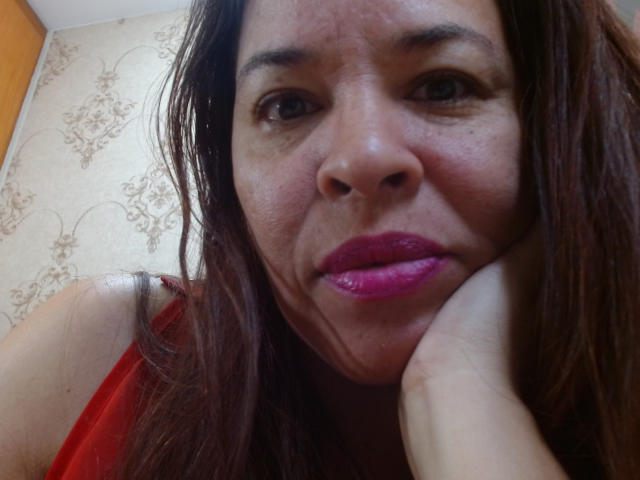 Fotografía de primer plano de LizethBells con cabello castaño largo, lápiz labial rosa y una blusa roja, apoyando su barbilla en la mano, mirando mirando directamente a la cámara con una expresión ligeramente seductora. El fondo está desenfocado..