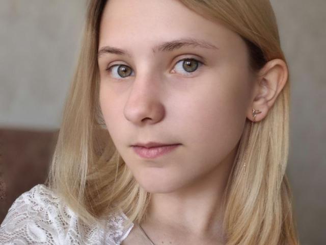 Close-up foto van RenaCloss met een lichte huid, blond haar en blauwe ogen. Ze heeft een neutrale uitdrukking, kleine, roze lippen, en draagt een witte kanten blouse. Subtiele seksuele spanning.