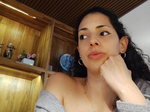 Foto ravvicinata di CurlyBlossom, una donna attraente con capelli scuri, grandi orecchini a cerchio e una maglia grigia a spalle scoperte, seduta. con il mento appoggiato sulla mano, lanciò uno sguardo languido e di lato. Mensole in legno sullo sfondo.