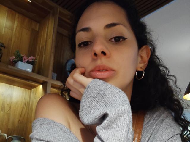 Close-up foto van CurlyBlossom, een vrouw met krullend zwart haar, die een grijze top met een open schouder draagt en haar hand op haar wang, kijkend naar de camera met lichtjes openstaande lippen. Houten planken op de achtergrond.