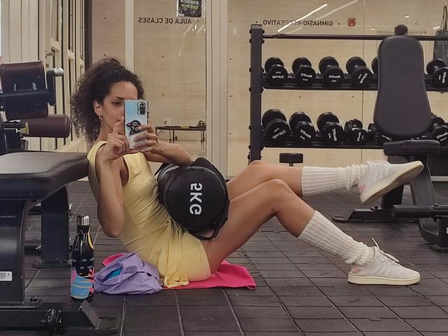 CurlyBlossom, mit lockigen Haaren, trägt ein gelbes Sportkleid und weiße Socken und sitzt auf einer pinken Matte in einem Fitnessstudio und hält ein ein schwarzes Gewicht im Schoß, macht ein Selfie.