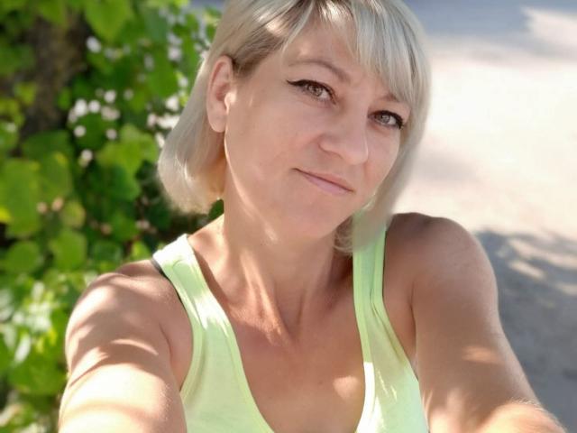 Selfie de primer plano de YasmineXHot, rubia, con un corte de pelo bob corto, vistiendo una camiseta verde neón. La luz del sol ilumina su rostro, verde. Fondo con follaje, ligera sonrisa, sombras suaves, ambiente informal, entorno al aire libre..