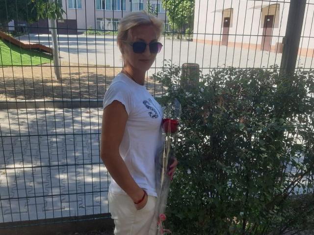 YasmineXHot, seitlich vor einem Drahtzaun stehend, trägt ein weißes T-Shirt und eine weiße Hose und hält ein rotes Getränk. mit kurzen, blonden Haaren und einer Sonnenbrille, in einer koketten, selbstbewussten Pose..