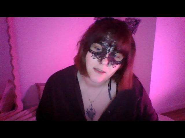 KittyTheWomen trägt eine schwarze Katzenmaske aus Spitze mit Katzenohren, ein schwarzes Oberteil und eine Halskette und sitzt vor einem rosafarben beleuchteten Hintergrund. Der Gesichtsausdruck ist leicht offenmundig, was sexuelle Spannung erzeugt. Weiches Licht.