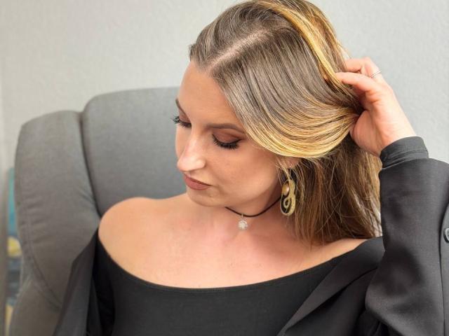 Milagres, with fair skin and long blonde hair, touches her head while looking down. She wears a black off-shoulder top, gold hoop earrings, and a small necklace. Fucking sexy.