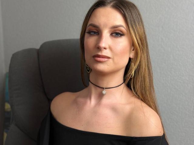 Milagres tiene el pelo largo y castaño, ojos azules, y lleva una blusa negra con un corte que deja los hombros al descubierto, junto con un collar negro. Está sentada, mirando directamente a la cámara, con una ligera sonrisa. Increíblemente atractivo/a..