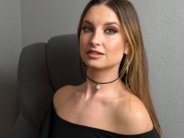 Milagres has long brown hair, blue eyes, and a slight smirk. She's wearing an off-shoulder black top, a black choker with a star pendant. Sexual tension, attractive, direct gaze.