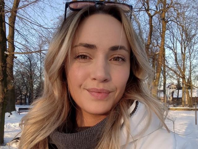 Close-up van ScarlettLips met lichtbruin, golvend haar, die een witte jas draagt over een grijze coltrui. Ze glimlacht, met een zonnebril op haar hoofd. Een met sneeuw bedekte park in de achtergrond..