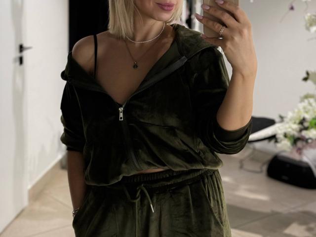 RoseGloss, blonde bob, takes a mirror selfie, wearing a black off-shoulder top with a visible zipper, black pants, and a necklace. Her hand covers part of her face, lips slightly parted.