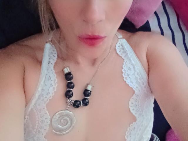 Primo piano della parte superiore del corpo di Maytena, con particolare attenzione al suo décolleté, che indossa un reggiseno di pizzo bianco. Indossa una collana di perline nere con un ciondolo a spirale d'argento. Le sue labbra sono leggermente socchiuse.