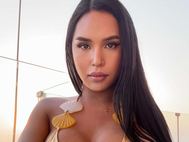 SavannaStallionCock has long, straight black hair, tan skin, and full lips. Wearing a gold shell necklace with yellow seashells covering her cleavage. Serious expression, subtle makeup, gold cross necklace.