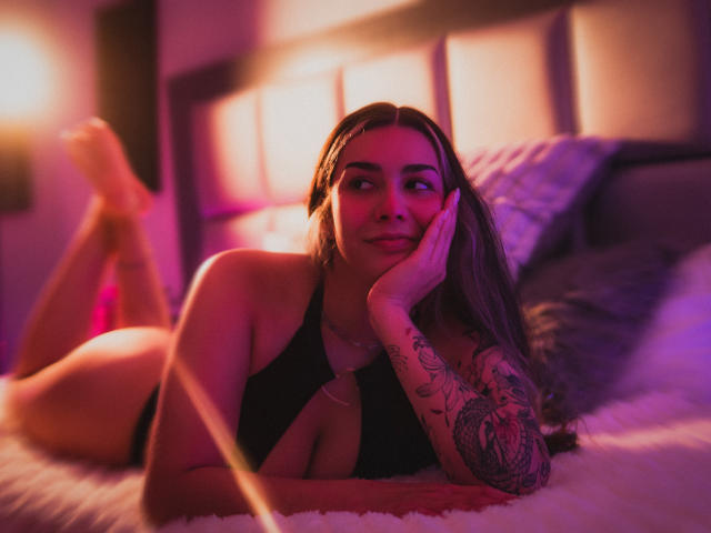BonnieRider ligt op een bed, badend in roze en paars licht. Ze draagt een zwarte top, heeft tatoeages op haar armen, leunt met haar kin op haar hand en heeft haar benen gebogen bij de knieën. Heel aantrekkelijk..