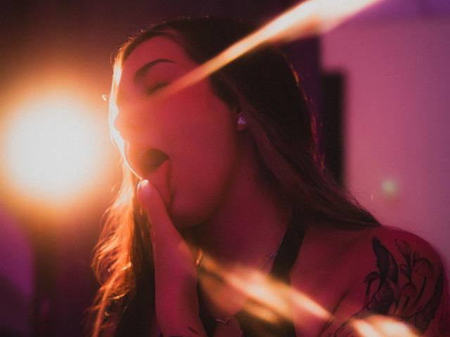 Photographie de BonnieRider, les yeux fermés, les lèvres légèrement entrouvertes, la main près du visage, une source de lumière vive illuminant la partie gauche. côté, effets de lumière rose et orange, tatouage sur l'épaule droite, expression suggestive.