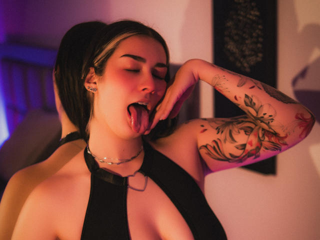 BonnieRider, tätowiert und oben ohne, zeigt die Zunge und legt eine Hand an die Wange, trägt ein schwarzes Halstuch, mit dezenten rosa und violetten Farbtönen. Beleuchtung, kurze schwarze Haare, verführerischer Ausdruck, Armtattoos, erotische Spannung.