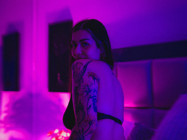 Photographie de BonnieRider, une femme tatouée avec de longs cheveux noirs, un soutien-gorge noir, le dos tourné, regardant par-dessus son épaule, baignée dans lumière néon violette et rose. Tension sexuelle, tatouage visible sur l'épaule, regard séduisant.