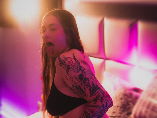BonnieRider, uma mulher de cabelos longos com intrincadas tatuagens nos braços, língua de fora, vestindo uma blusa preta, com iluminação em tons de rosa neon e laranja. Características do fundo: cama desfocada com travesseiros. Tensão sexual, pose provocativa, expressão sedutora..