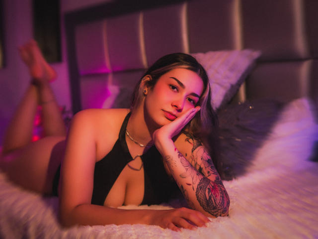 BonnieRider está acostada en una cama con la cabeza apoyada en su mano, vistiendo un top de tirantes negros que muestra un brazo tatuado. La iluminación rosa y púrpura crea un brillo seductor. Increíblemente sexy..