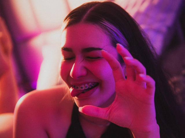 Fotografía de primer plano de BonnieRider, una joven con cabello largo y oscuro, sonriendo con los ojos cerrados, con la mano cerca de la cara, de color rosa y iluminación neón púrpura.
sus dientes son visibles, su expresión es coqueta..