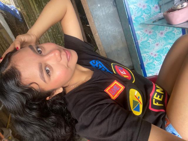 JuliaBennett with black hair, tan skin, and red nails, sitting with one arm behind her head, wearing a black sports shirt with colorful logos, intense gaze, sexual tension, outdoor setting.
