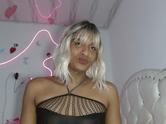 Photograph of IdiiaLatina with bleached blonde hair and bangs, wearing a black mesh top that reveals her small breasts, pouting with full lips. Neon lights and a white headboard in background.