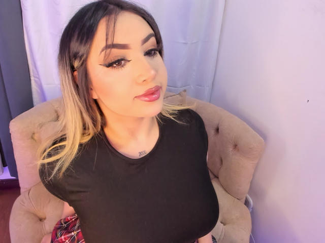 MelissaLust, sitting in a beige tufted chair, wearing a black top, with long black and blonde hair, heavy eyeliner, and red lipstick, looking at the camera with a slight smile.