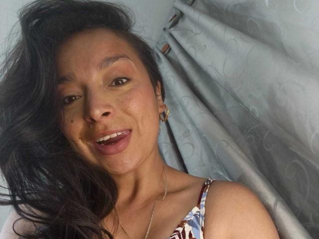 Close-up of StangelarRivera with dark, wavy hair, smiling with slightly open mouth, brown eyes, wearing a floral top, light blue curtains in background, soft natural light, casual, sexy vibe.