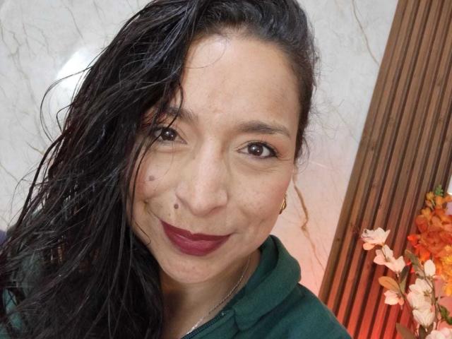 Close-up of StangelarRivera, a woman with wet black hair, dark red lipstick, and a nose piercing, smiling subtly. She's wearing a dark green hoodie. Orange flowers in the background.