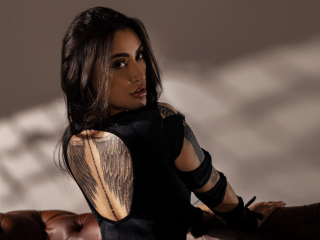 LiliValeria mit dunklem Haar, die über ihre Schulter schaut, trägt ein schwarzes Oberteil mit einem aufwendigen Flügel-Tattoo auf ihrer rechten Seite. Schulter. Schattiges Licht, Ledersofa im Vordergrund. Verführerischer, intensiver Blick..
