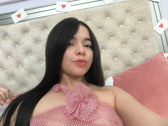 Lissy Carter, una mujer asiática con cabello largo y negro, lleva puesto un top rosa con tirantes y un lazo, y está acostada sobre un cabecero beige con textura. con los labios fruncidos y rosados, rodeada de pequeños corazones rojos.