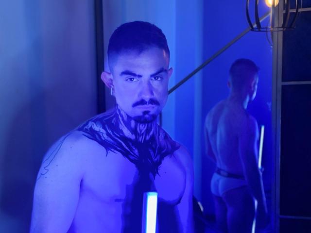 Fotografía de un hombre musculoso, con barba y cabello corto, con iluminación de tonos azules y un espejo reflectante en el fondo. DallasWhitee está sin camisa, vistiendo una bufanda negra con un aspecto brillante y húmedo. La luz azul crea tensión sexual..