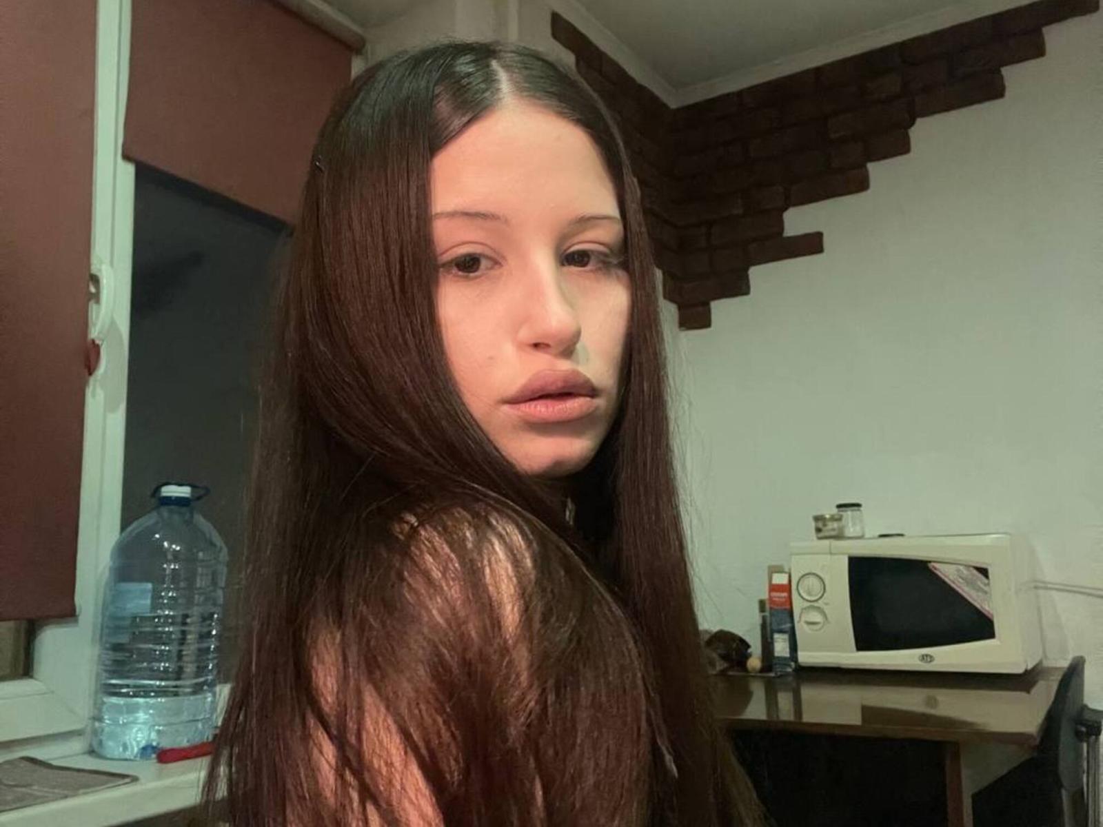 KimikoFranqui, with long straight black hair, gazes over her shoulder, light illuminating her face, sitting in a dim room with a water bottle and microwave on a desk. Fucking intense gaze.