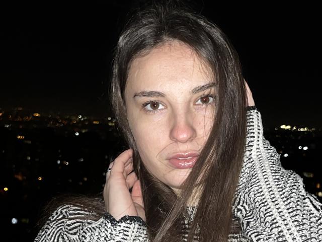Close-up photograph of JosephineRowe, a young woman with long dark hair, brown eyes, and glossy lips. She's wearing a zebra-print coat, her hand touching her neck. Dark background with city lights.