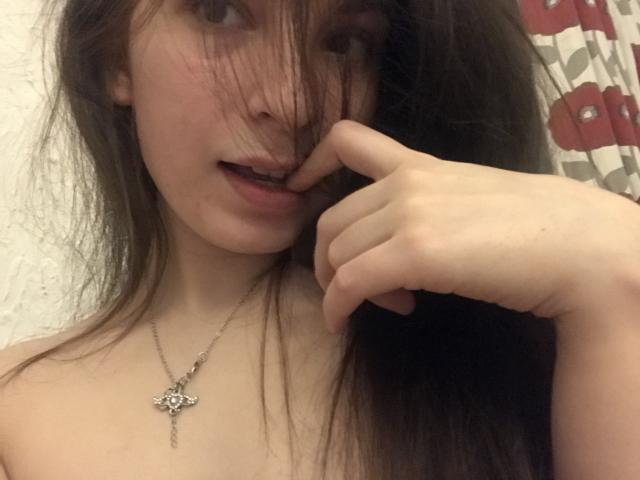 Photo en gros plan de JosephineRowe, à la peau claire et aux longs cheveux bruns, qui couvre partiellement son visage et mord sa propre doigt. Expression séduisante, petit collier en argent avec un pendentif en forme de fleur, l'arrière-plan présente un tissu à motifs rouges et blancs..