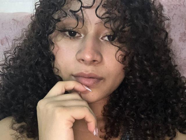 Close-up of SofiaPassionne with curly black hair, light brown skin, and full lips. She gazes directly at the camera, her index finger touching her lower lip, creating intense sexual tension.