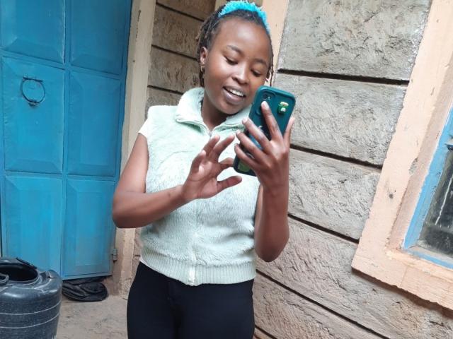 RachieChristina with dark skin and blue braided hair, smiling, holding a green phone, wearing a white sleeveless top, black pants, standing in front of a blue door and wooden wall.