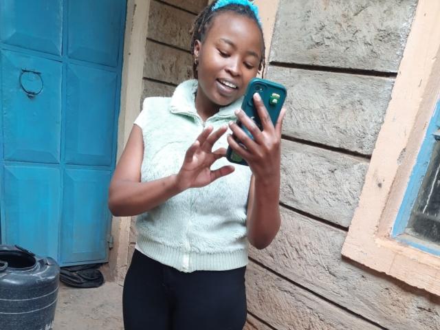 RachieChristina steht draußen, lächelt und macht ein Selfie mit einem grünen Handy. Sie trägt ein weißes Top ohne Ärmel und schwarze Hosen, ein blaues Haarreif, hält ein Telefon mit der rechten Hand und sieht verführerisch aus..