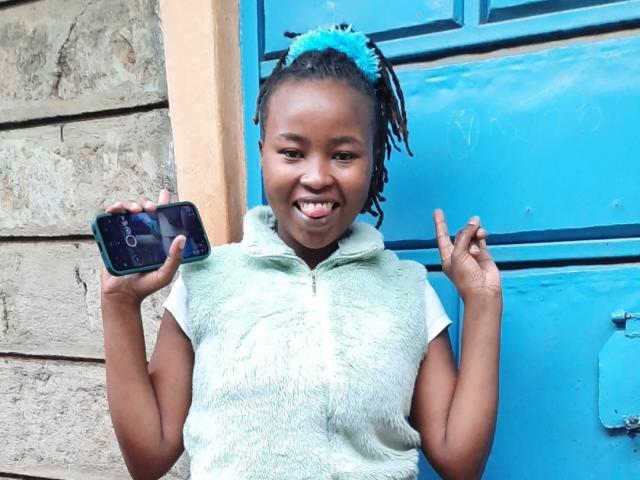 RachieChristina with dark skin and blue hair ties, grinning widely, holds a black phone in her right hand, flashing two fingers on her left. Wearing a white textured shirt, blue door behind.