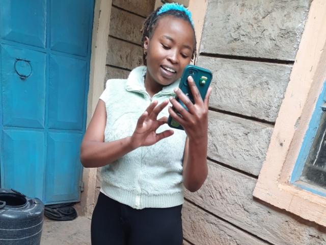 RachieChristina with dark skin and braided hair, wearing a white sleeveless top and black pants, holding a green phone, smiling, standing against a blue door and beige wall. Fucking hot.