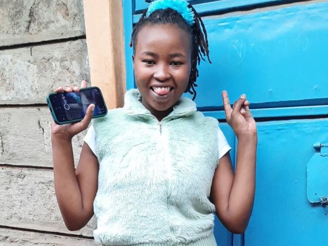 RachieChristina with dark skin and braided hair, wearing a white textured shirt, holds a black camera in one hand and flashes a double peace sign with the other, smiling widely. Bright blue door in background.