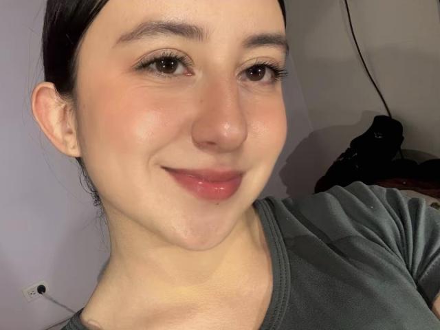 Close-up selfie of RosalieDupont with pale skin, dark hair pulled back, brown eyes, and a subtle smile. She wears a dark gray shirt, slightly flushed face, and natural makeup. Background blurred.
