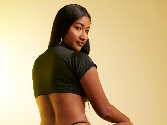 Fotografia de Aainoha, uma mulher de pele escura com cabelos longos e pretos, olhando por cima do ombro, vestindo uma cropped preta justa. parte superior, mostrando suas costas, fundo amarelo dourado. Corpo incrivelmente atraente e bem definido..