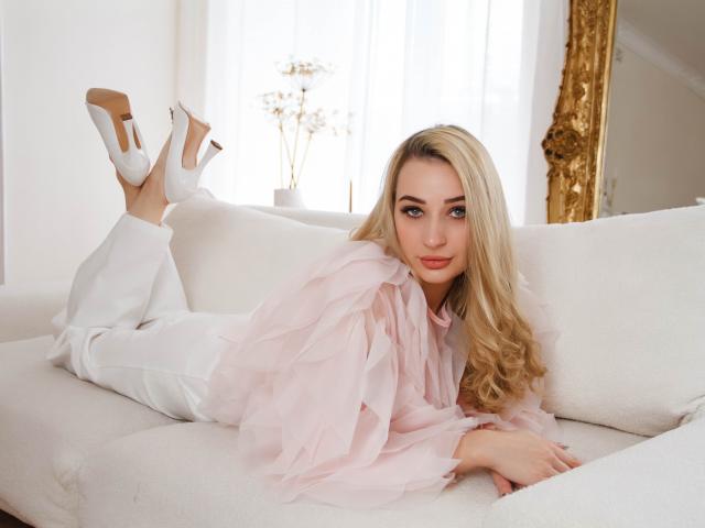 BarbaraRosy, a blonde woman with long hair, lies on a white couch, legs up, wearing a pink tulle top and white pants, white heels, seductive gaze, bright light background, gold frame.