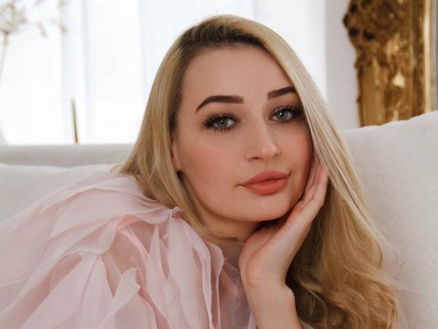 BarbaraRosy with blonde hair, blue eyes, and pink lips, wearing a sheer pink top, resting her face on her hand, looking directly at the camera with a sultry expression. Soft lighting.